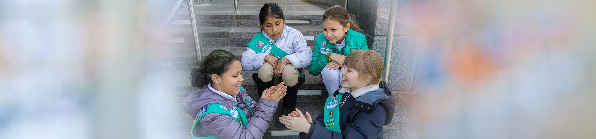  Group of four Junior Girl Scouts sitting on some granite steps. Two of them are playing a hand game while the other two watch. 